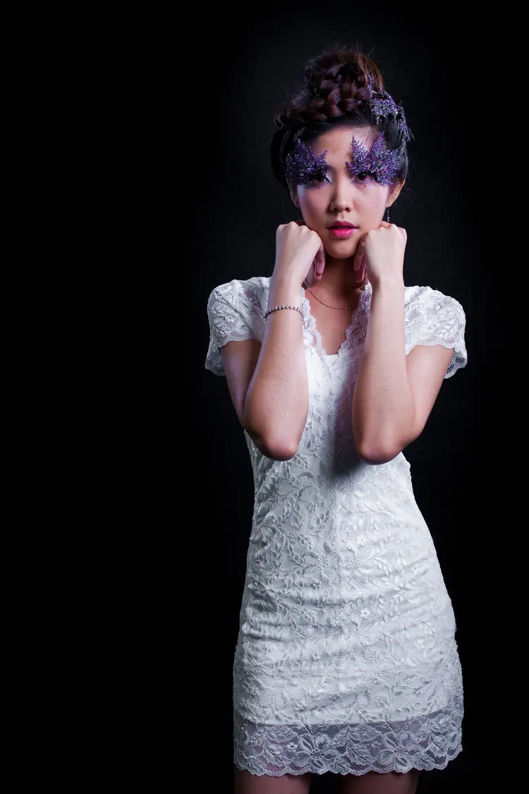 A woman in a white lace dress with purple eye make-up, lit against black.