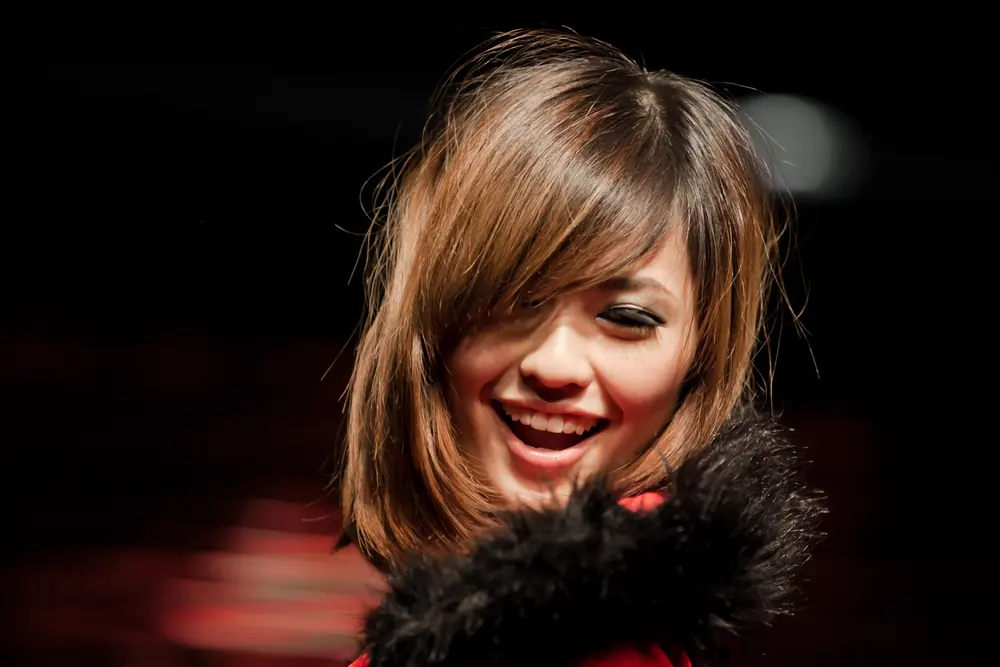 A woman laughs with her hair across her face, lit against a black background.