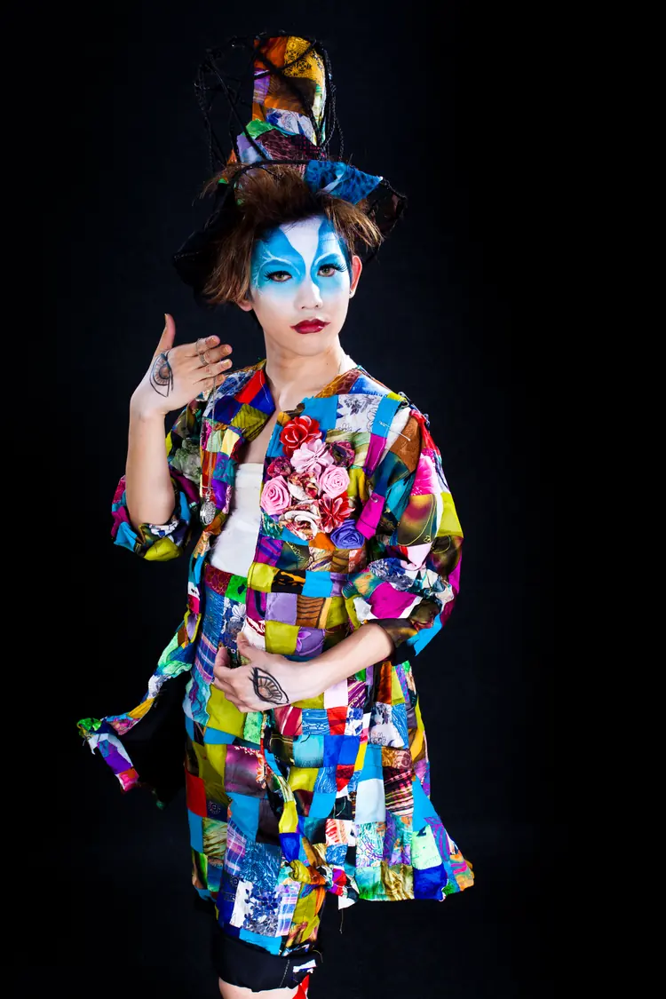 A model with blue face paint and a patchwork coat, lit against black.