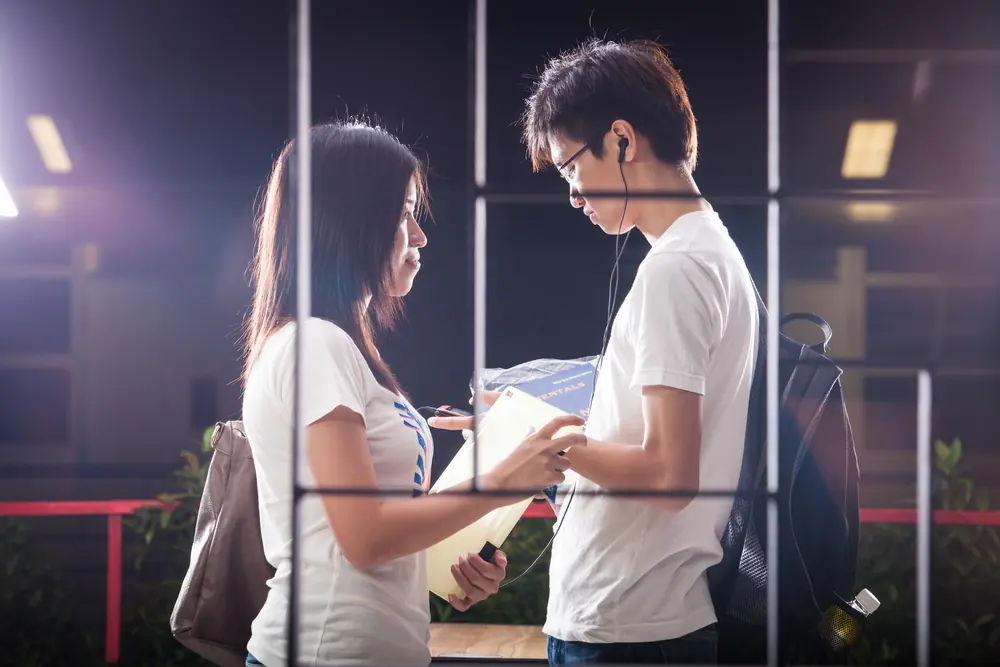 Two young people look at notes behind a window frame, in purple evening light.