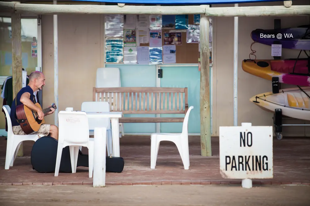 A man plays guitar outside a beach kiosk, next to a No Parking sign.