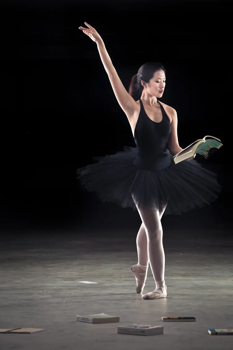 A ballerina on pointe reads a book on a dark stage scattered with books.