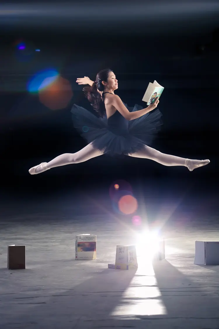 A ballerina leaps in mid-air holding a book, with a bright light behind her.