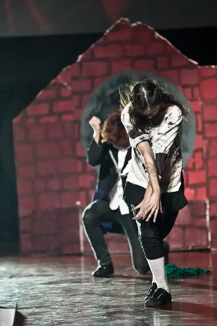 Two dancers perform in front of a painted brick house on stage.