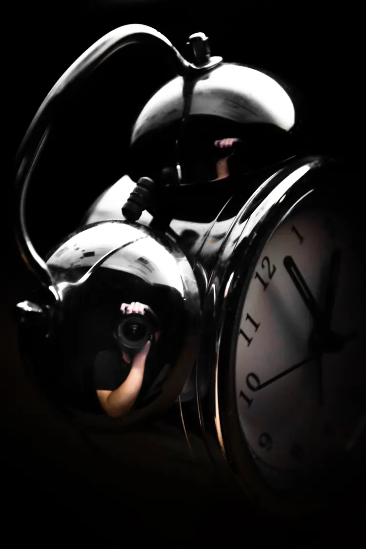 A chrome alarm clock against black. The photographer is reflected in its bells.